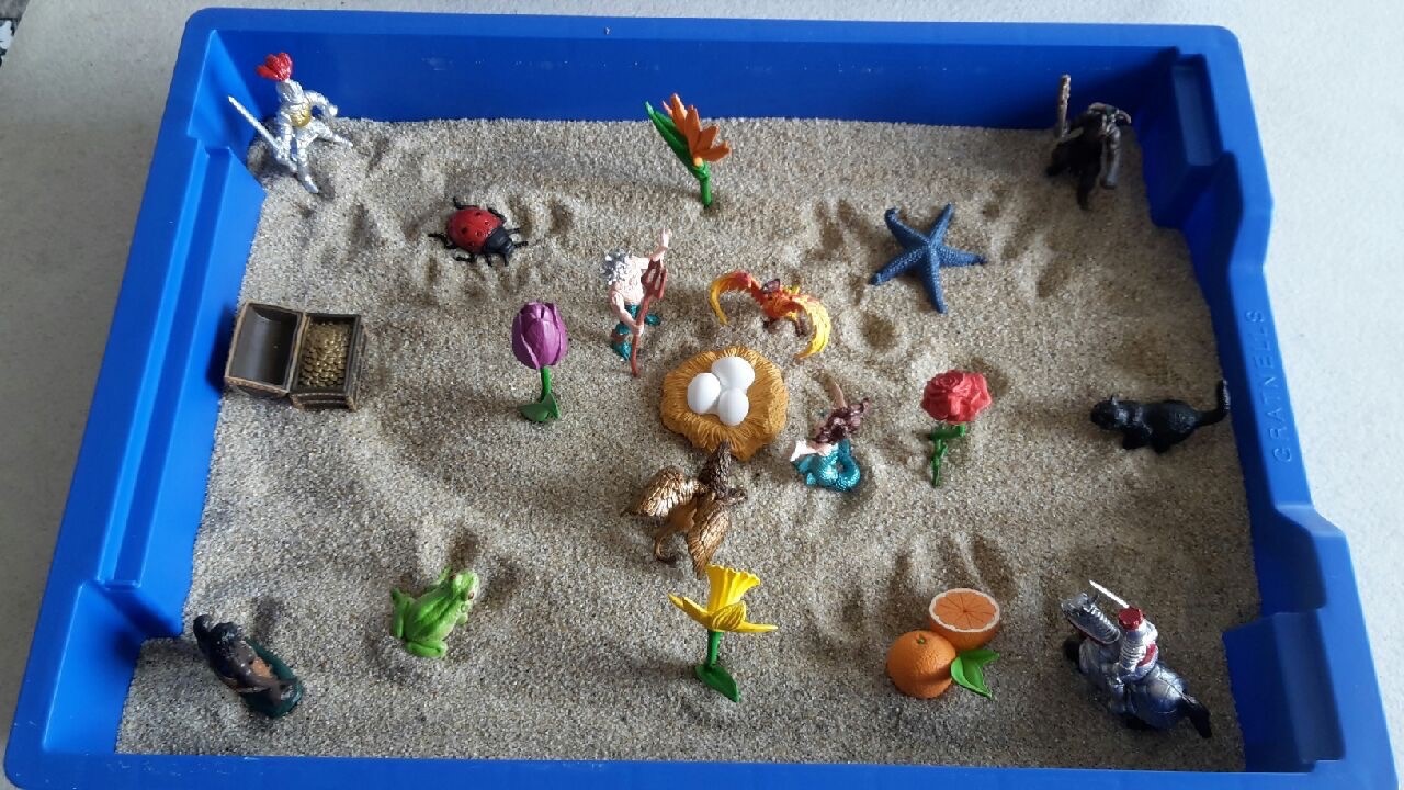 Legetøjsfigurer og genstande i en blå sandkasse med sand.